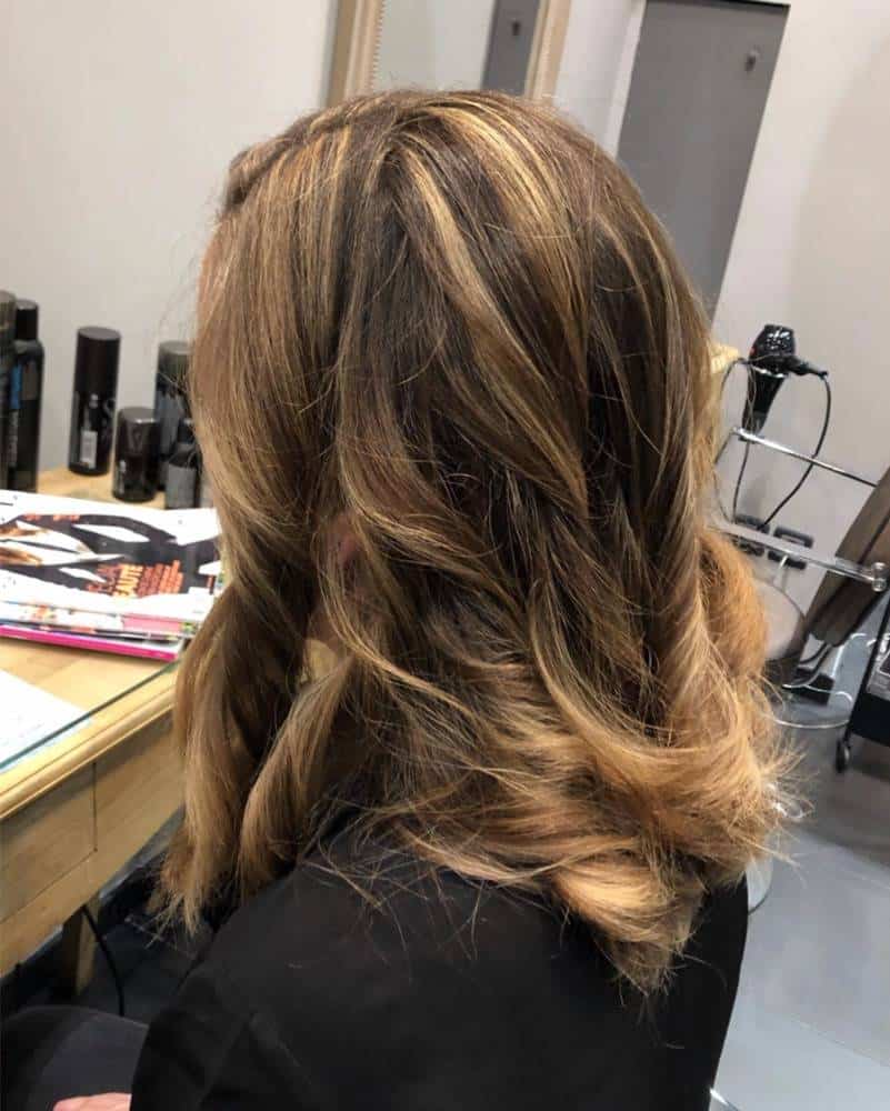 LE NUANCIER 4 Cheveux mi-longs ondulés avec des reflets blonds, vue de dos dans un salon de coiffure avec des produits et une chaise de coiffure en arrière-plan.